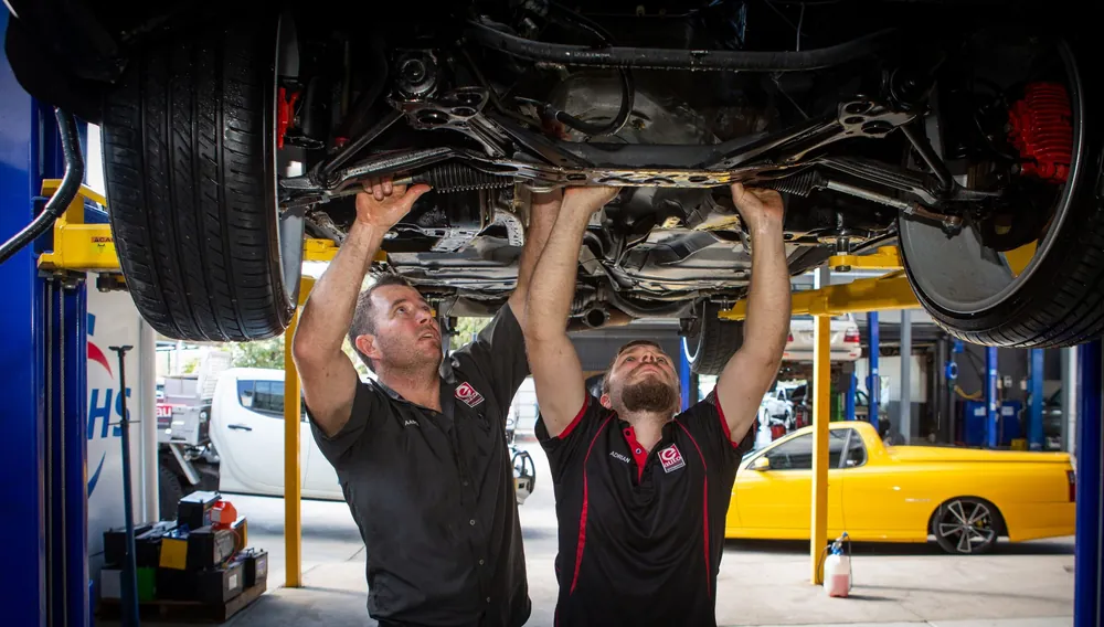 Two mechanics working under a raised car on a hoist in the Woree workshop